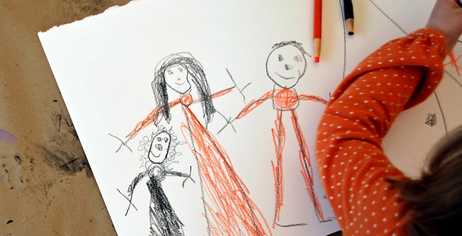 A child drawing three people on white paper using orange and black drawing tools.