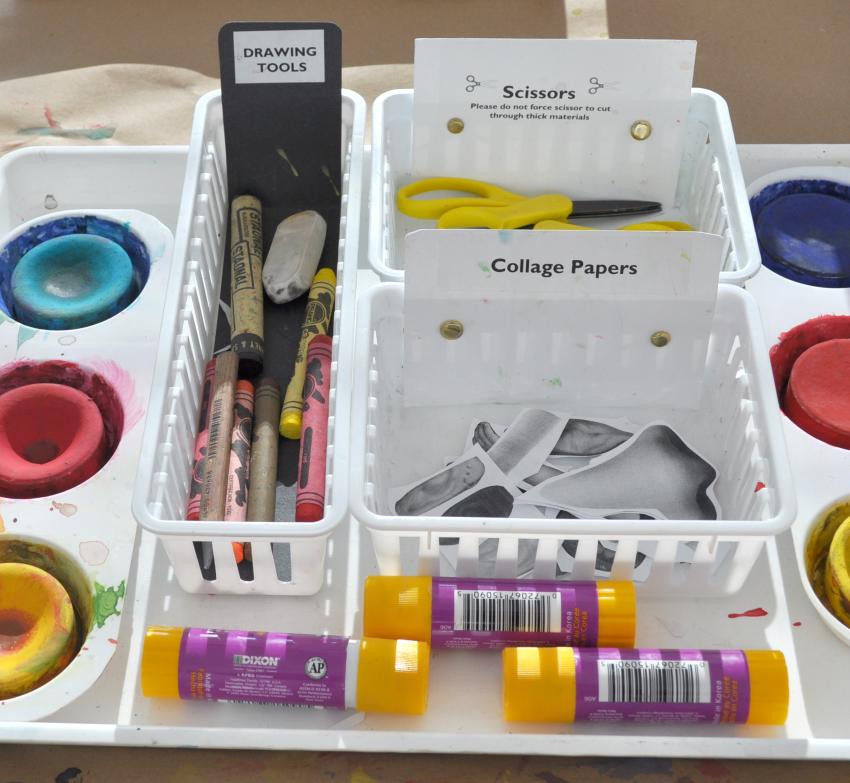 Several baskets with art supplies in them including scissors, collage papers, and watercolor paints.