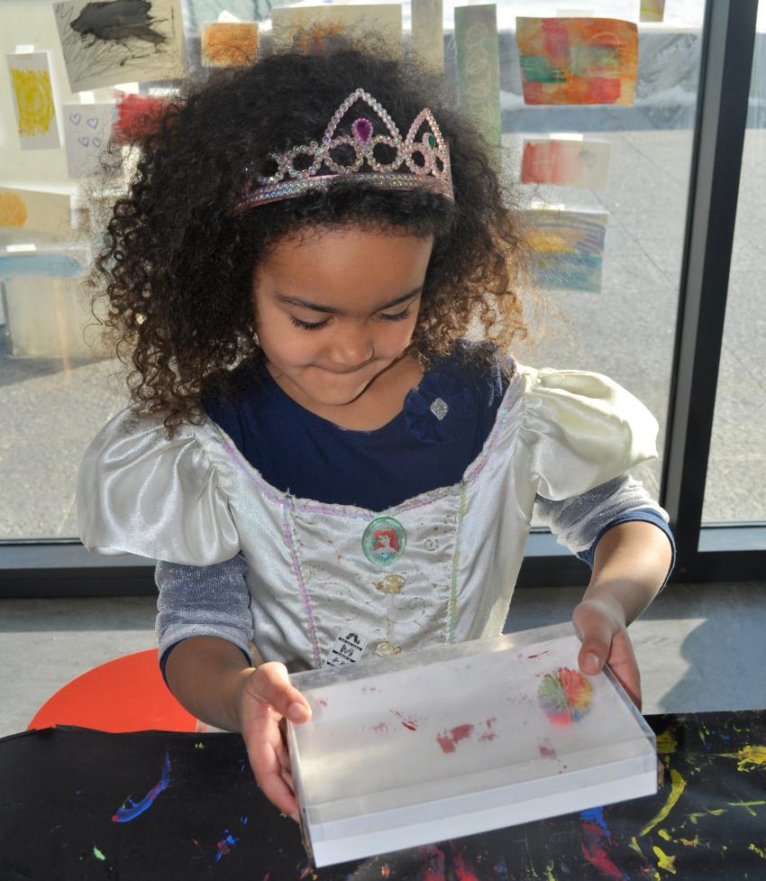 A young child wearing a tiara shakes a box with a painted ball inside.