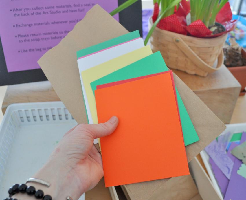 An adult holds a stack of colorful blank cards and a small brown paper bag.
