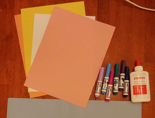 An array of materials including warm colored construction paper, markers, and glue. 