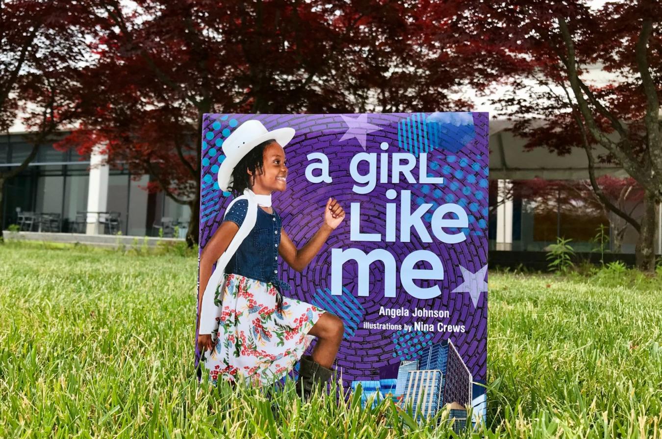 The book A Girl Like Me is propped in the green grass with trees in the background.