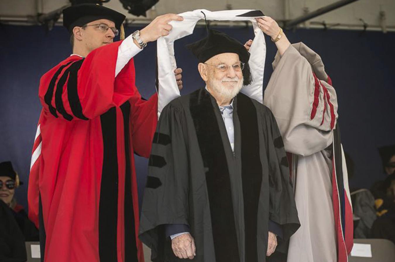 Eric Carle wearing black cap and gown with two people in red and gray gowns putting a white honorary degree hood over his head