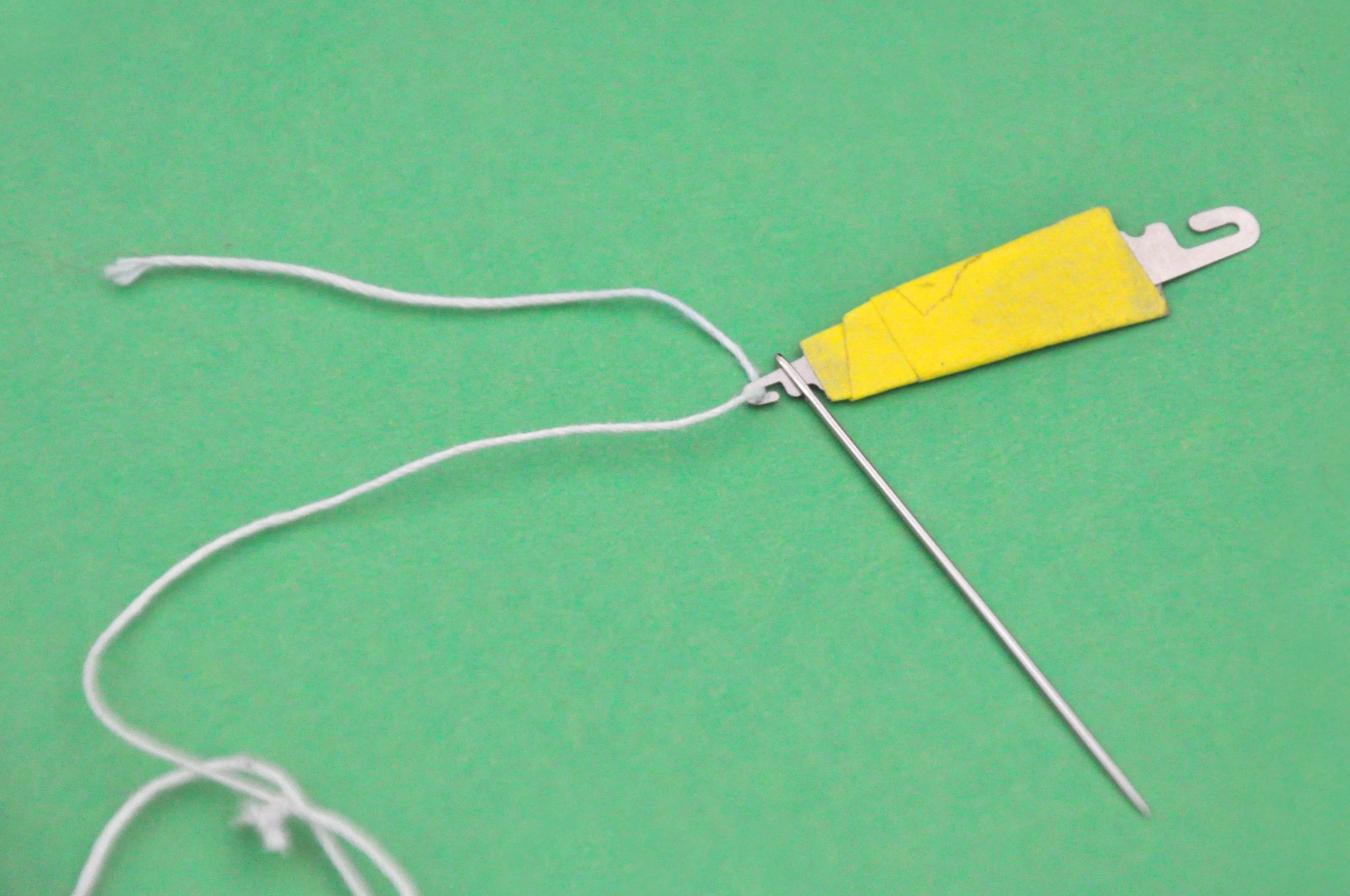 A needle threader, with a white thread on the hook, being put through the eye of a sewing needle.