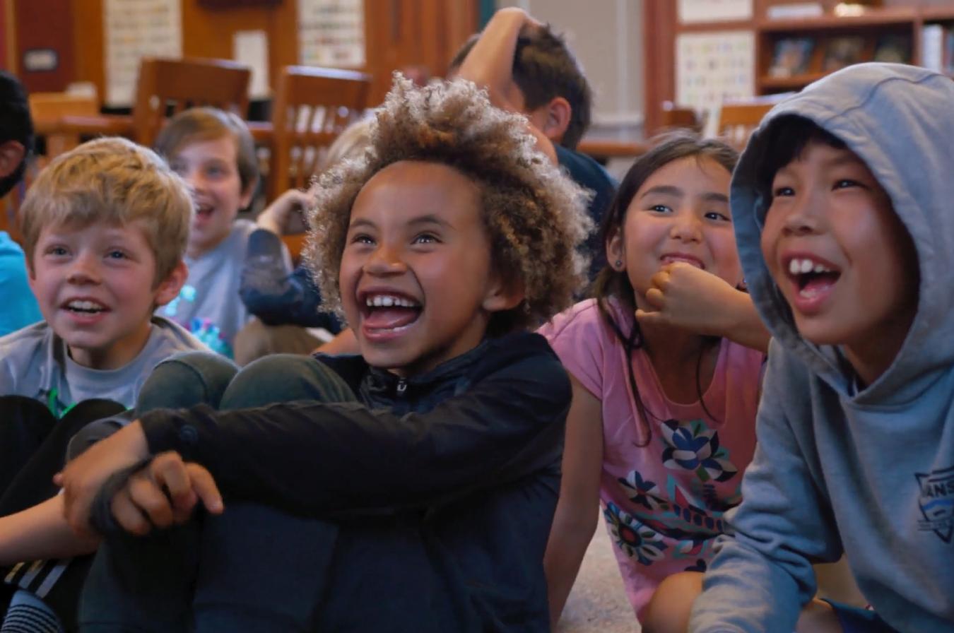 A group of children have big smiles on their faces as they sit on the floor of a school library listening to a story being read aloud by picture book maker Mac Barnett.