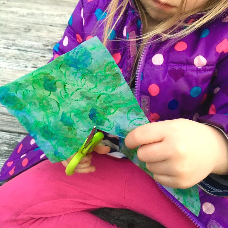 A young child cutting a piece of the watercolor-painted papers with scissors.