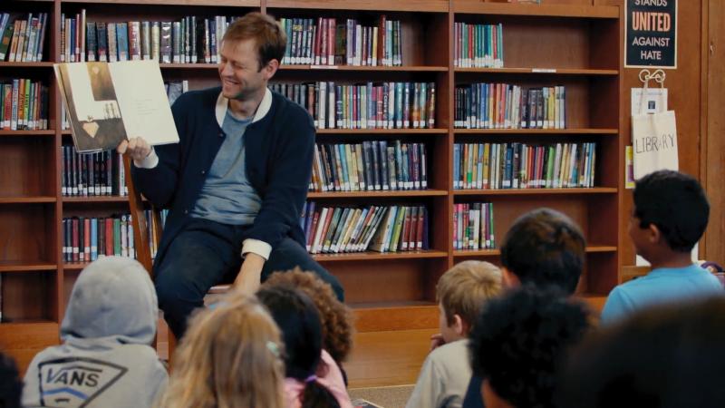 Mac Barnett, a children’s picture book author, laughs as he reads aloud from his book “Sam and Dave Dig a Hole” to a group of children at an elementary school.