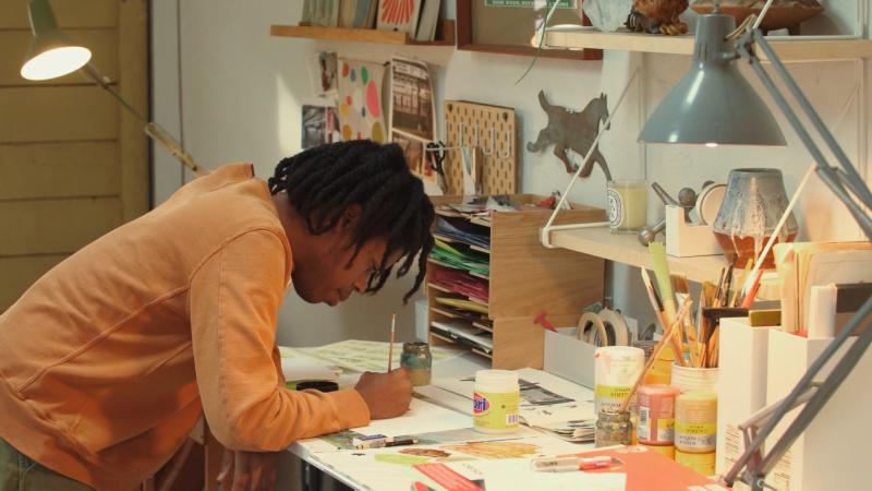 Children’s picture book author and illustrator Christian Robinson paints at his desk in a workshop filled with art supplies and illustrations.