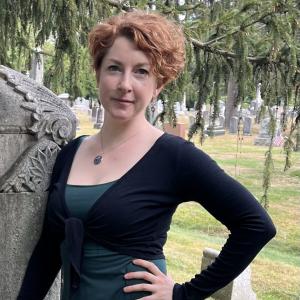 Woman standing against a rock with short curly hair, green dress and black cardigan