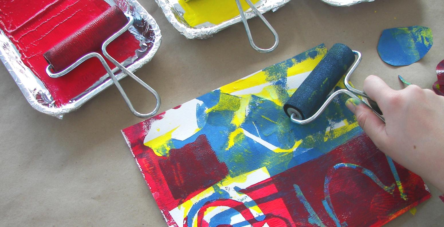 A hand using a brayer to roll red, yellow, and blue paint over stencils, creating a layered print.