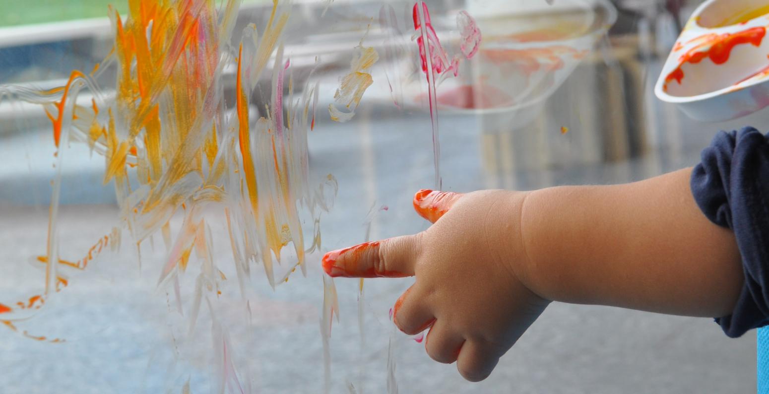 A small hand finger paints with warm colors on a window.