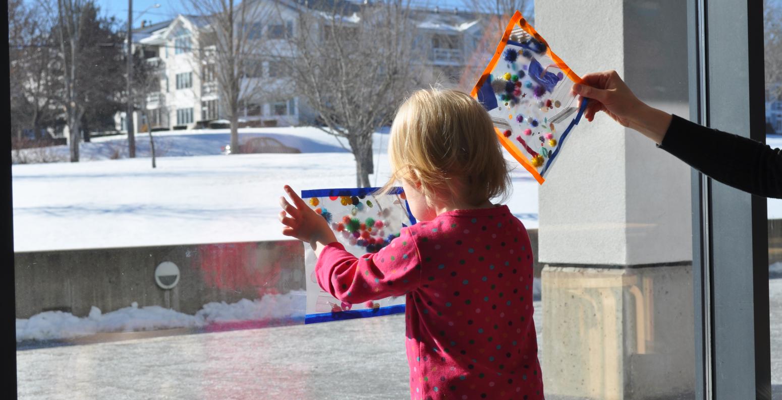 A young child holding their sparkly collage up to the window.
