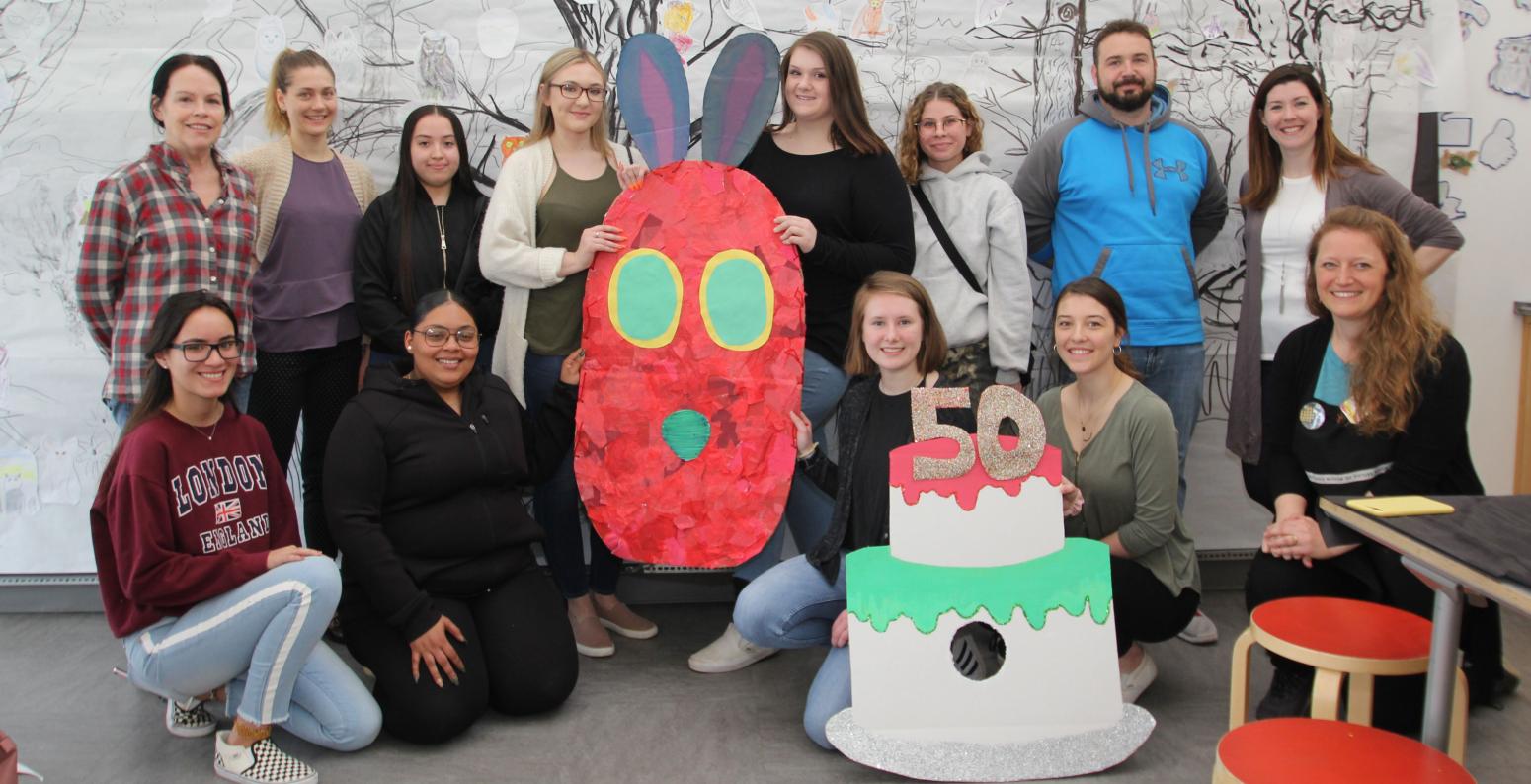 A group of HCC students and educators holding a 50th birthday cake and caterpillar head both made from cardboard and paper.