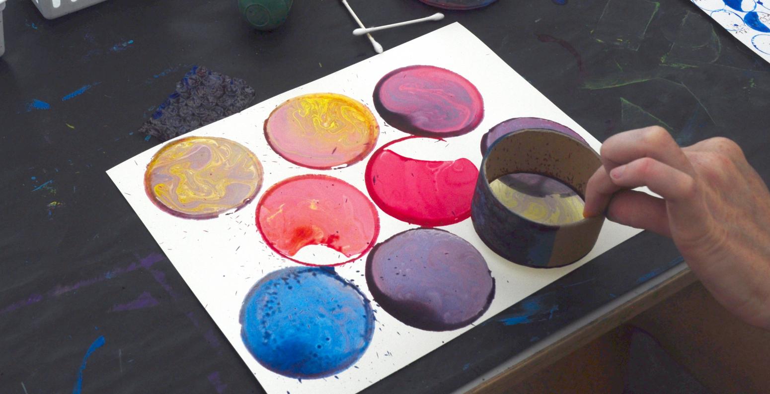 An adult prints circles onto white paper using a cardboard tape roll and tempera paint.