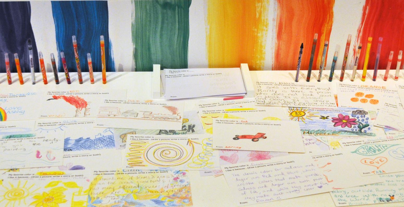 A table with the colors of the rainbow behind it, with colored pencils and postcards with drawings on them.