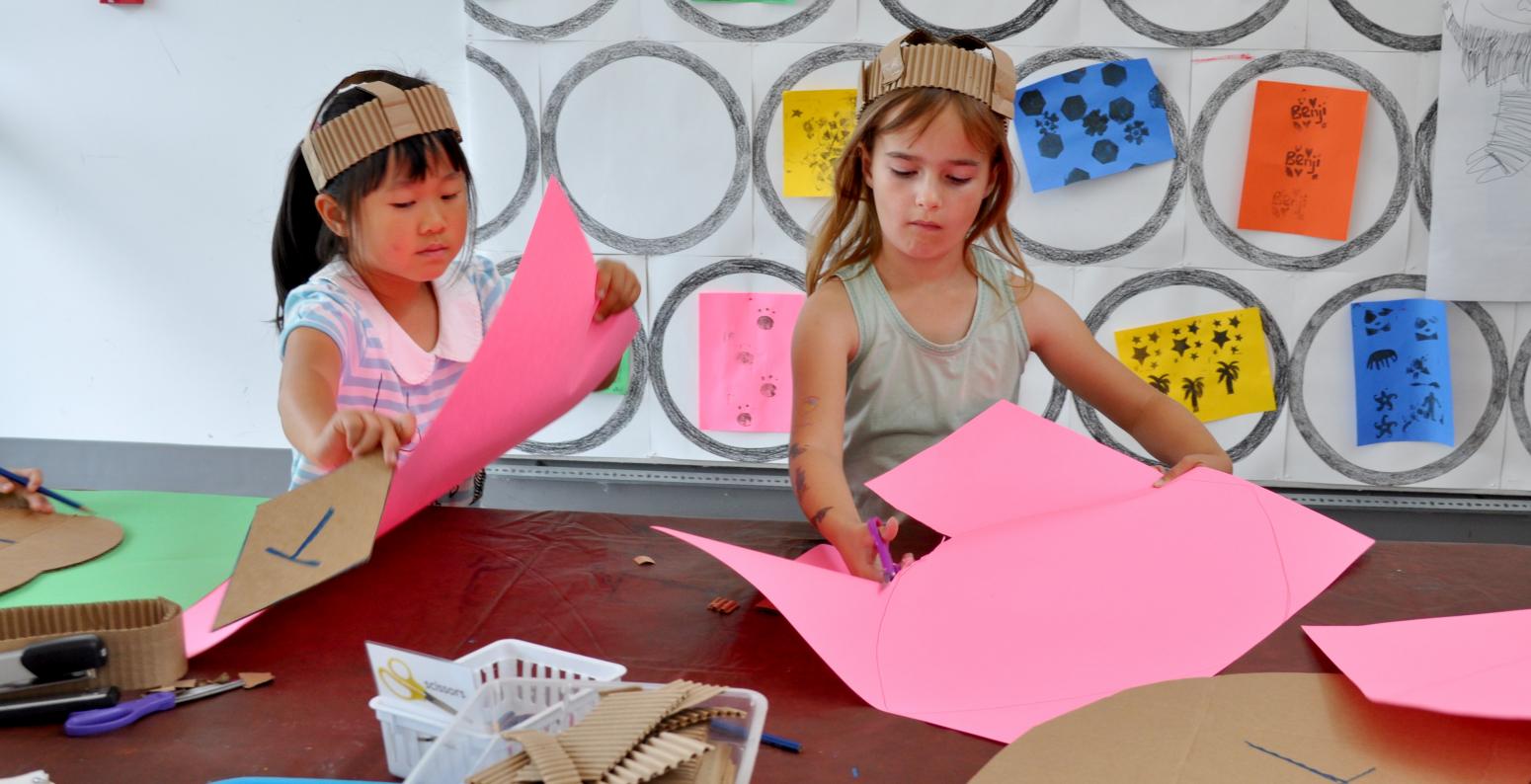 Two children making their own costumes, wearing cardboard hats and cutting pink paper.