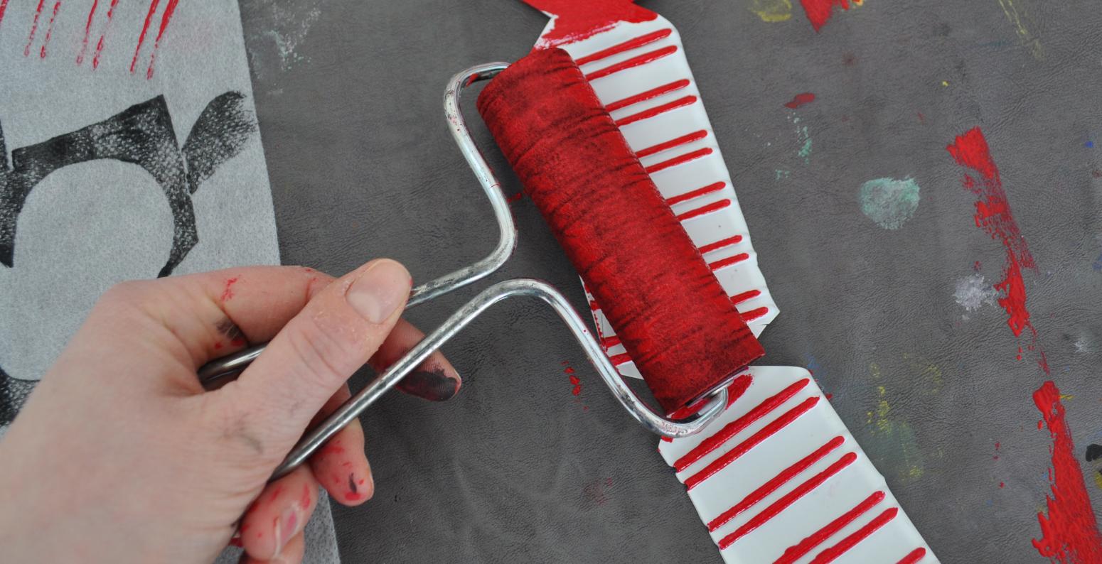 A hand applies red paint to a styrofoam packing material using a roller.