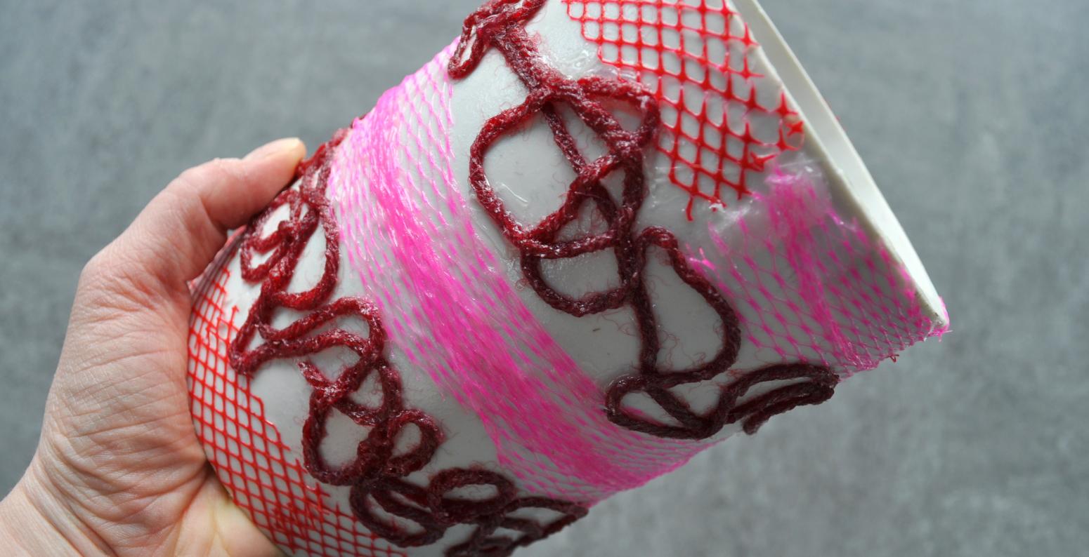 A hand holding a homemade printmaking tool, where a yarns and plastic mesh have been attached to a large PVC pipe.