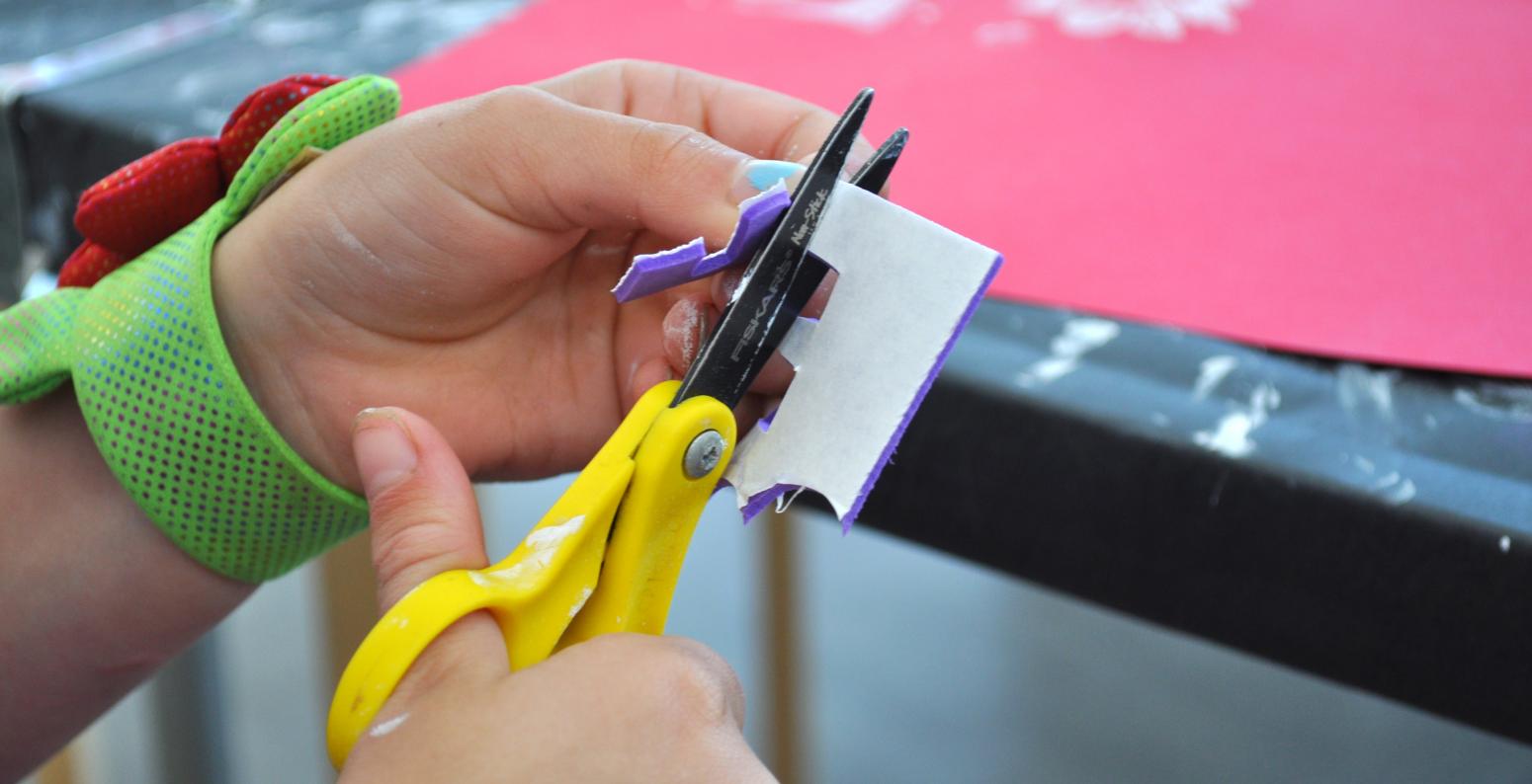 A hand cutting a piece of purple self-adhesive foam with a pair of yellow scissors.