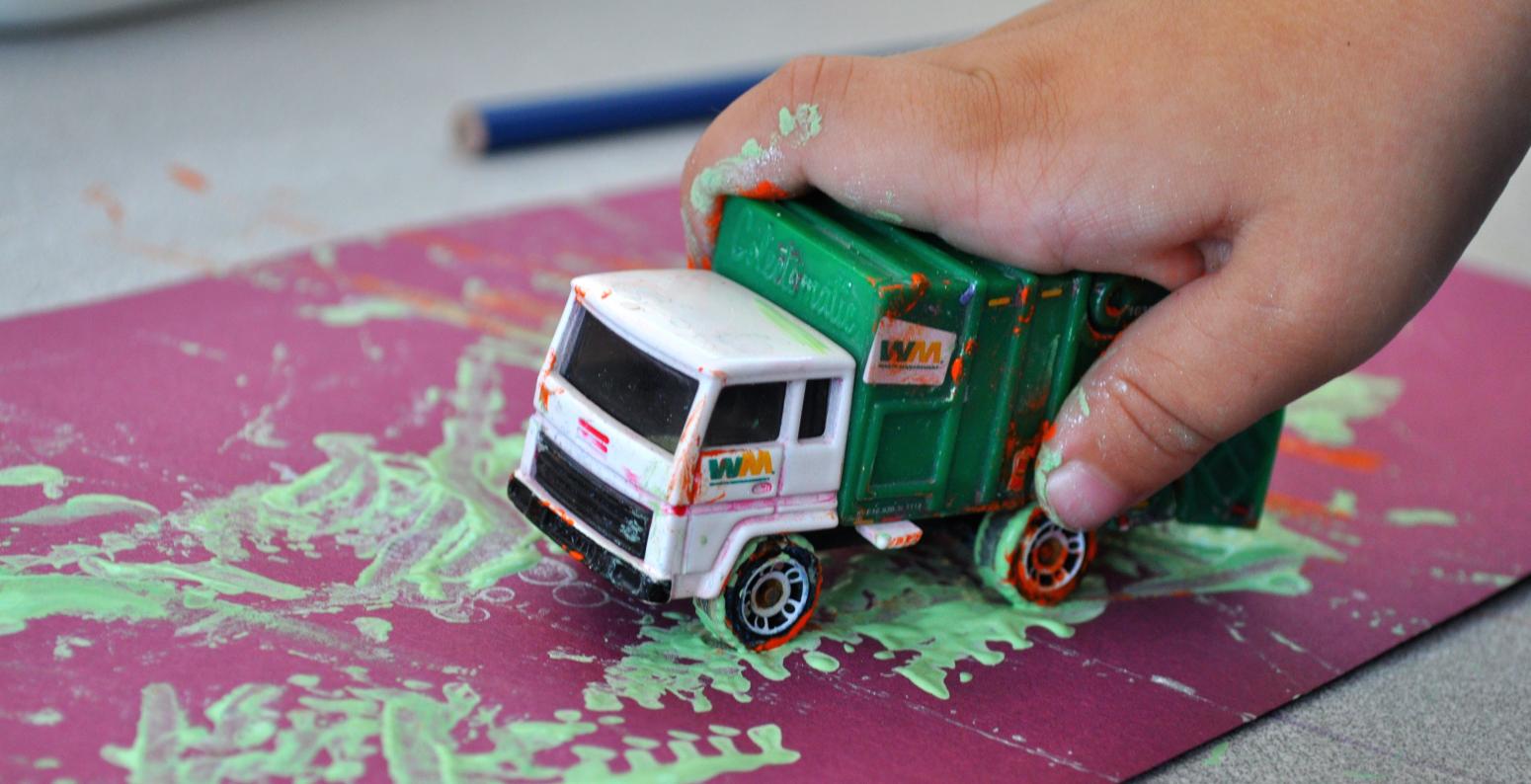 A young child's hand rolling a toy truck through green paint and onto a purple piece of paper.