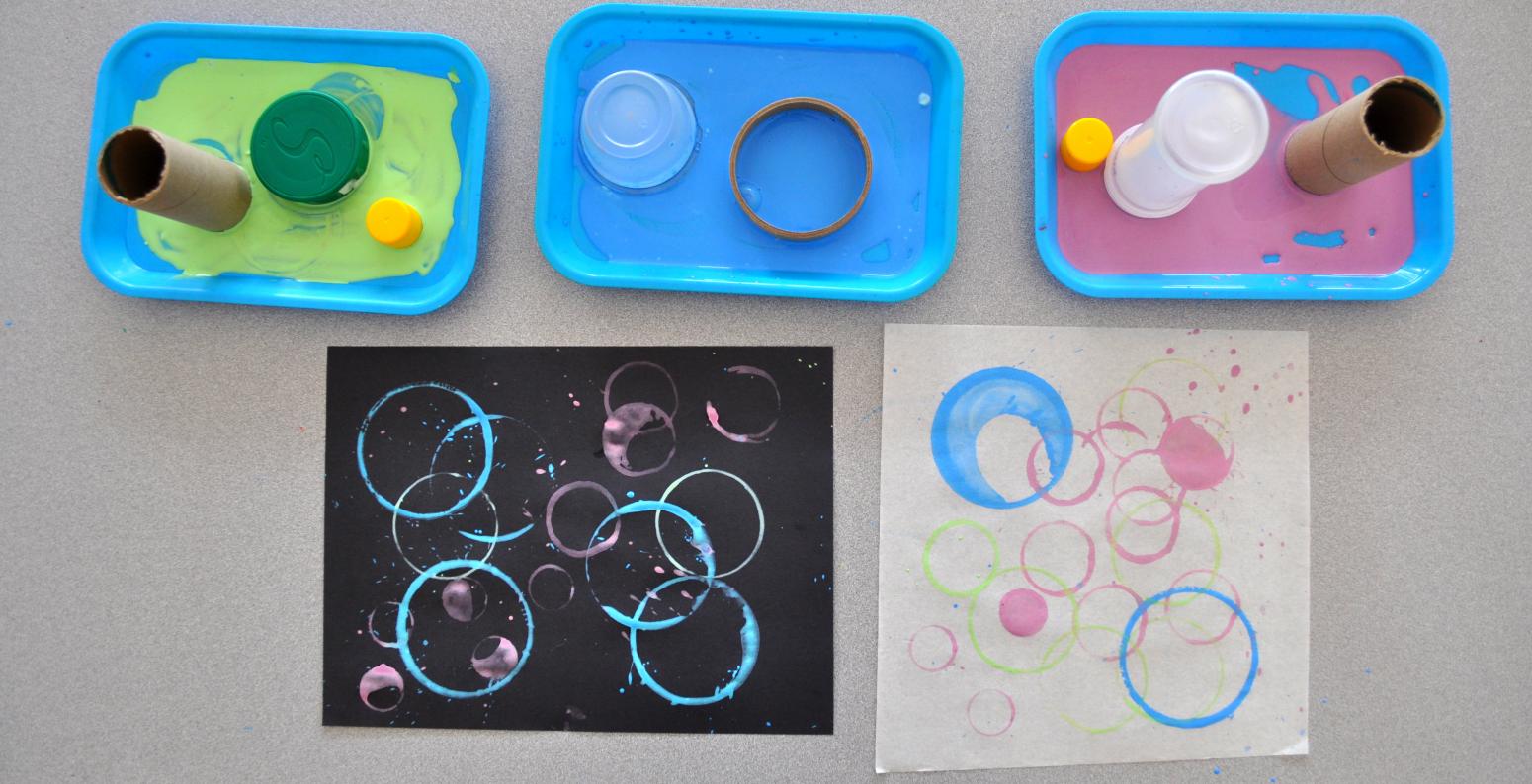 Three trays of different paint colors with circular objects on each, and two paintings with circle stamps on them.