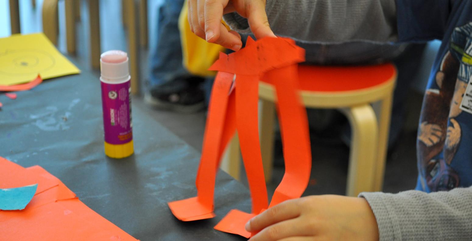 A young child's hand constructs a red elephant out of strips of paper, glue, and staples.