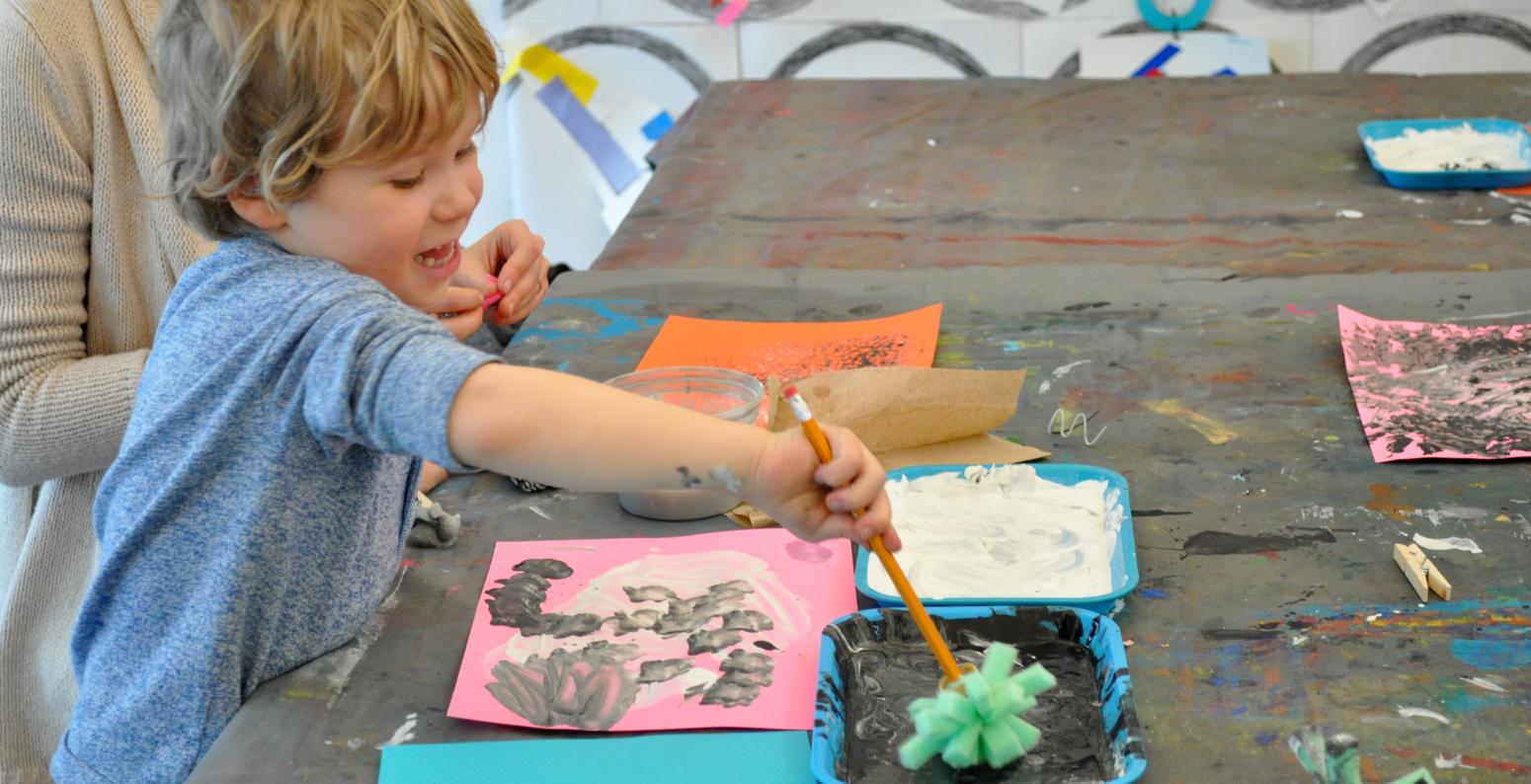 A young child enthusiastically painting with a textured paintbrush in black paint.
