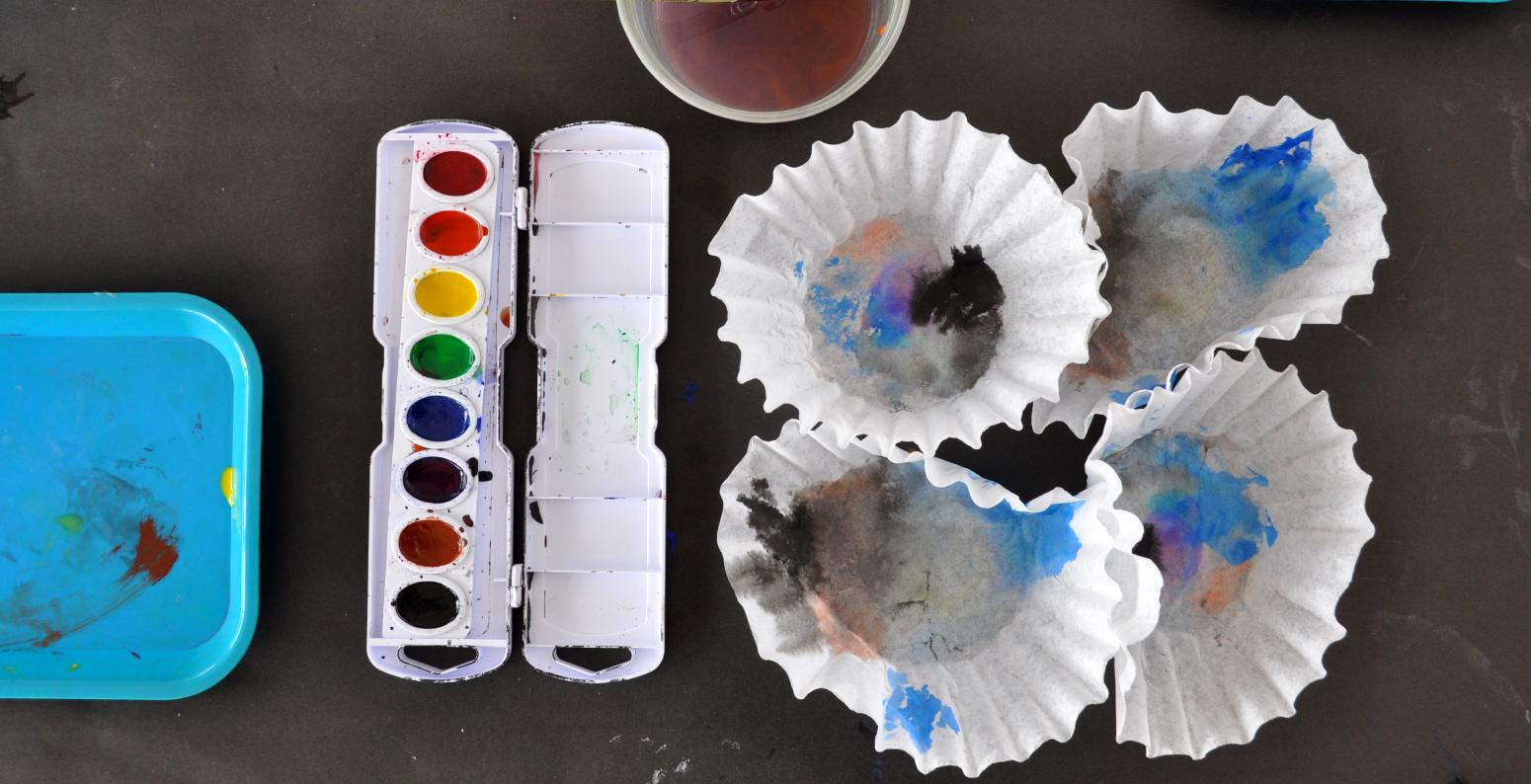 A watercolor paint set-up, with a palette, brush in water cup, and four coffee filters with paintings on them.