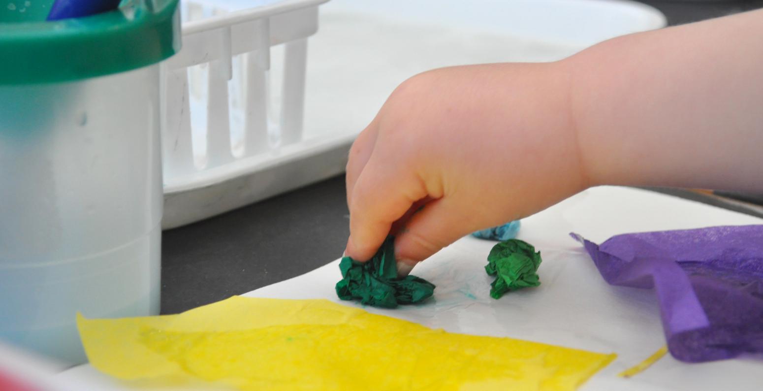 A young child gently places a crumpled piece of green tissue paper onto their background paper with glue.