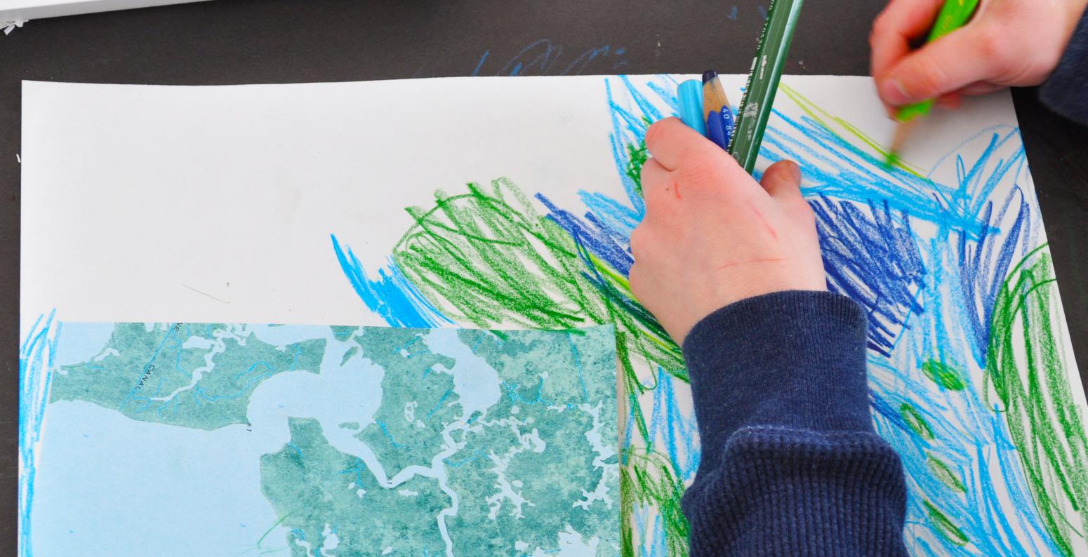 A young child draws with green and blue colored pencils to extend a piece of a map onto a larger piece of white paper.