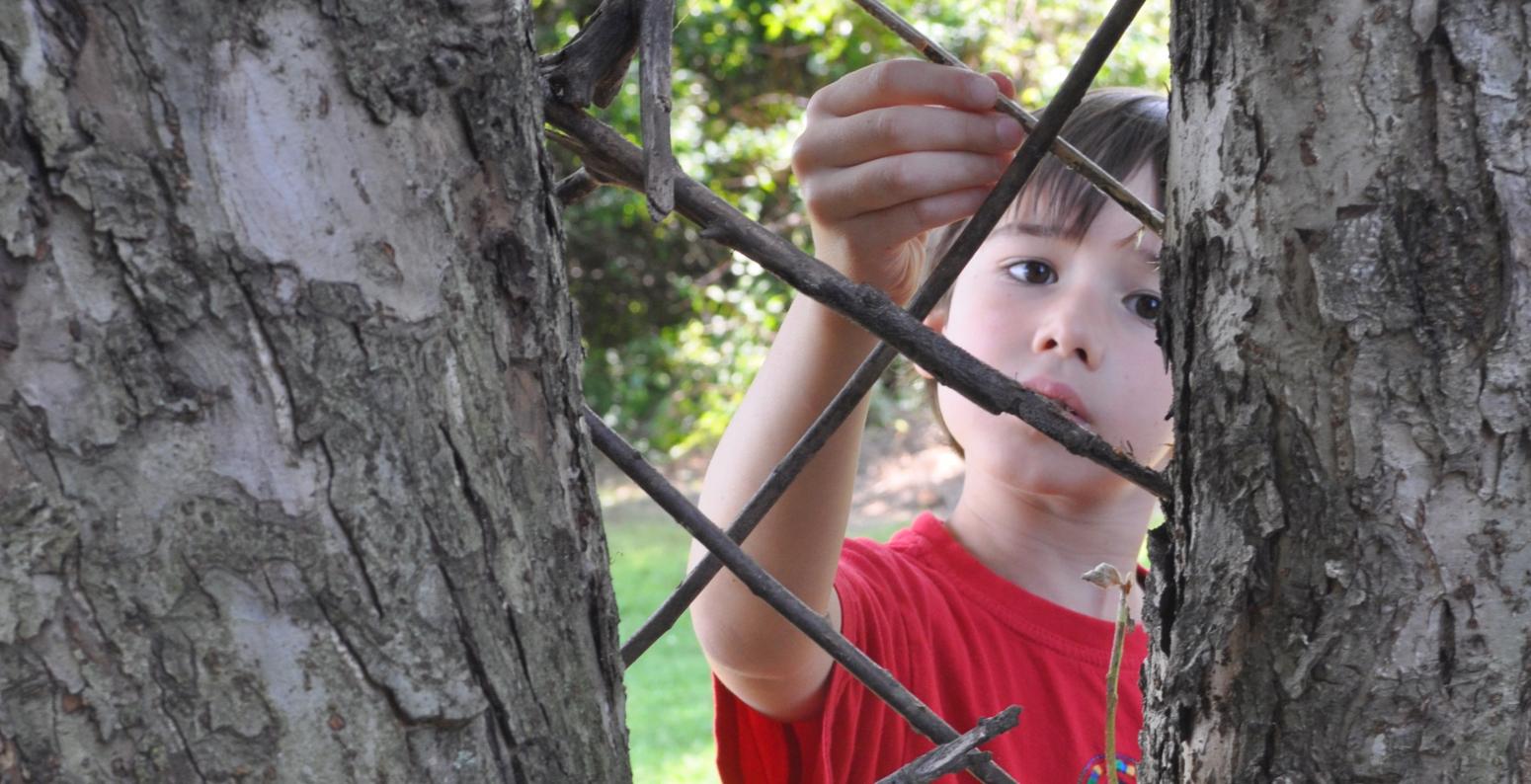 A child carefully places twigs between two tree trunks to create a diamond pattern sculpture.