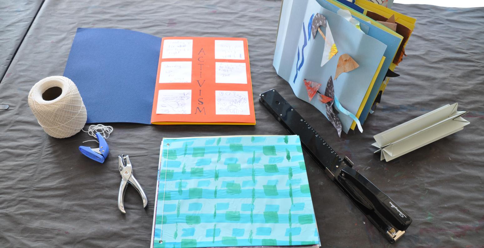 Three examples of books made from past Everyday Art Projects, accompanied by string, two-hole punchers, a crown binding, and a long reach stapler.  