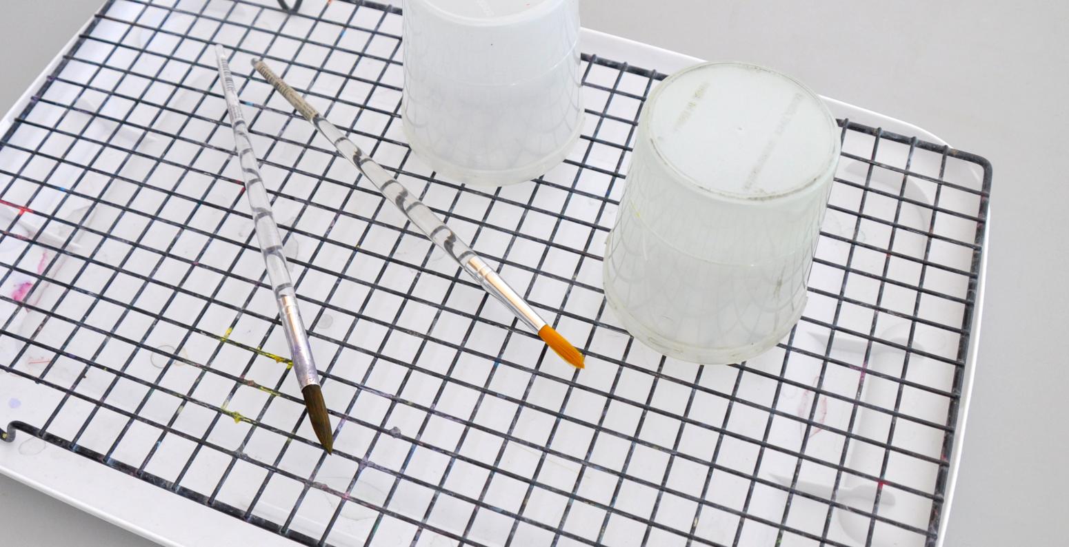 A drying rack made from a lid and a cookie drying sheet, with two brushes and two cups sitting on top drying.