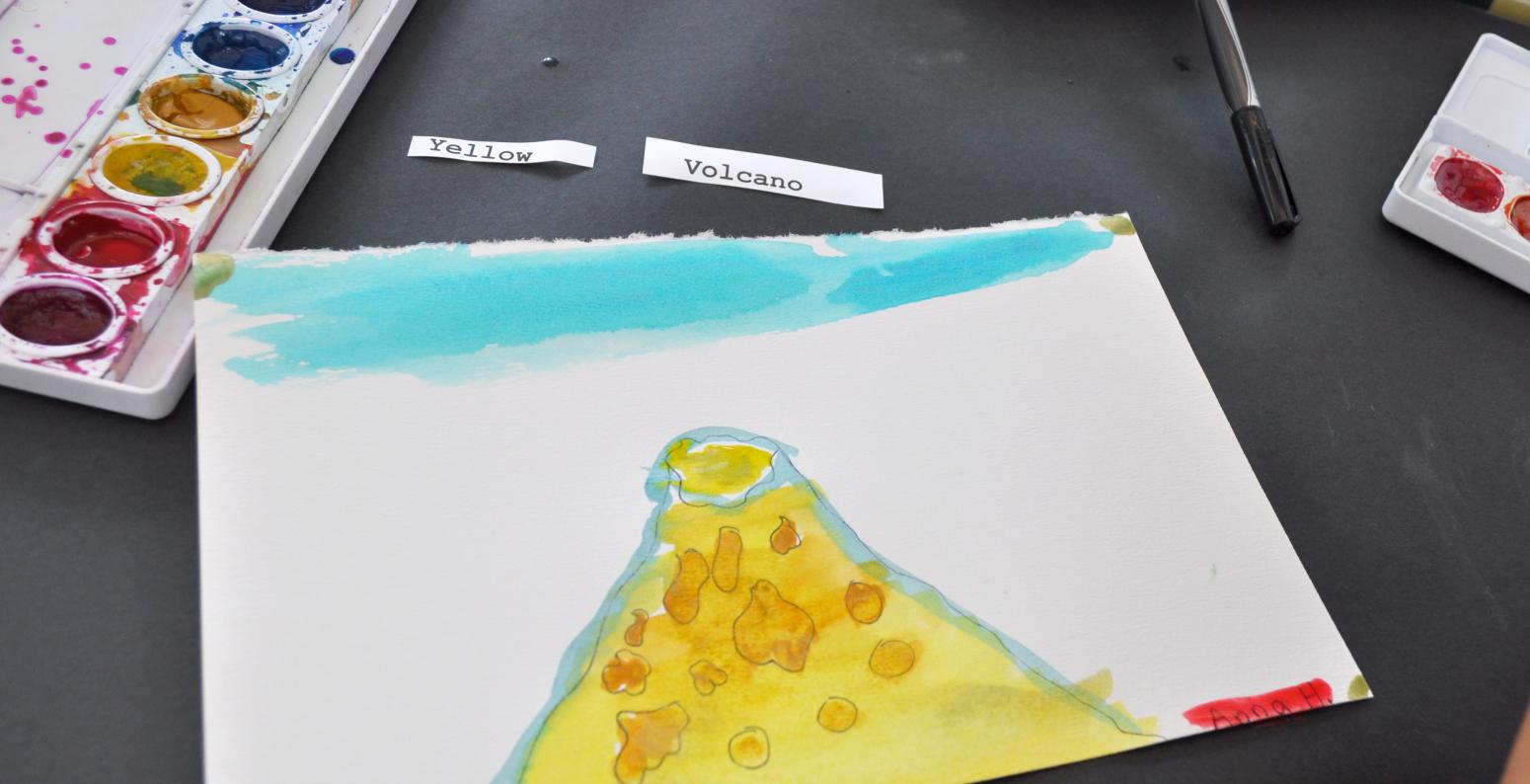 Two paper slips with the words "yellow" and "volcano" next to a watercolor painting of a yellow volcano against a blue sky.