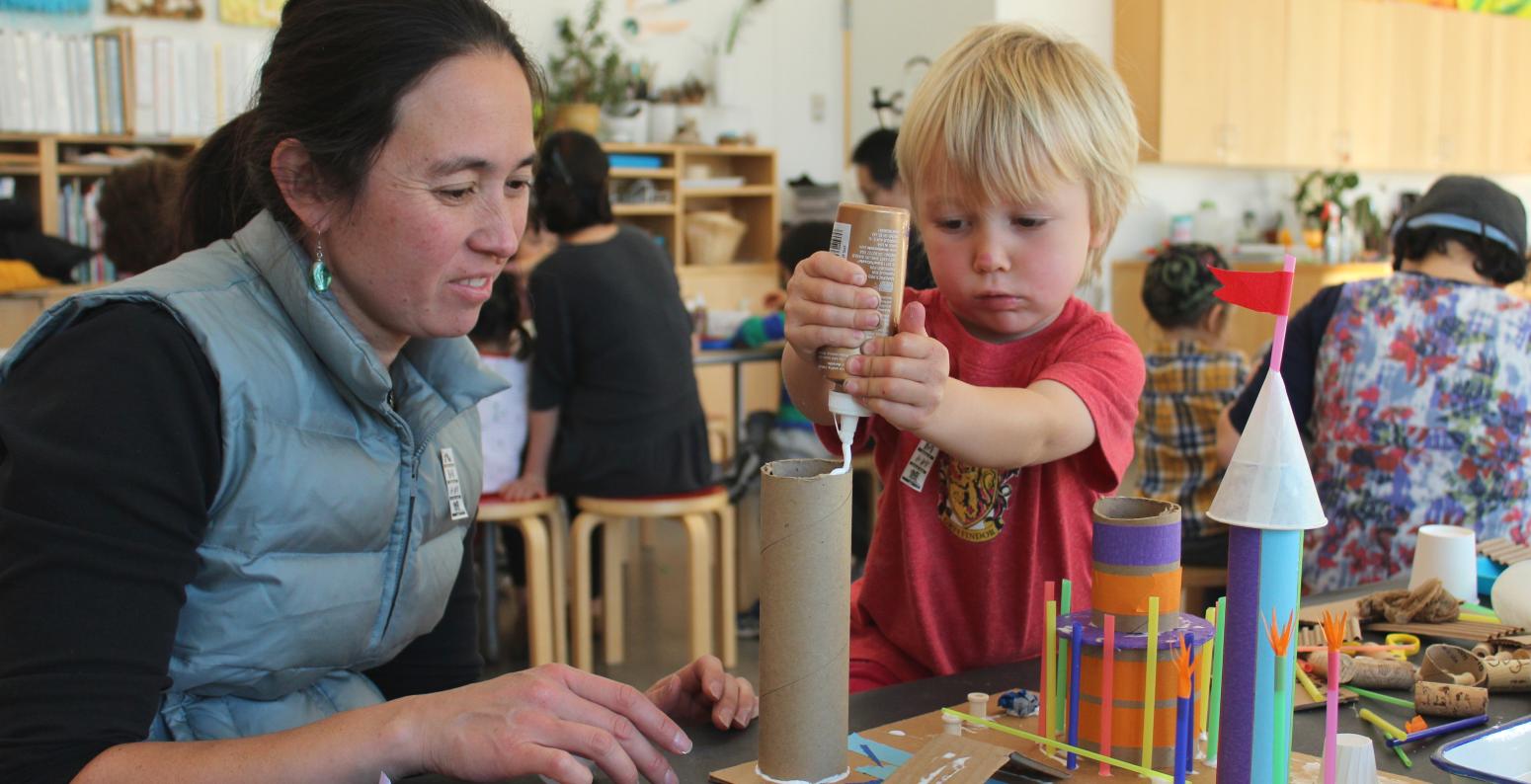 An adult and child work together to build a sculpture out of cardboard, glue, straws, and cups.