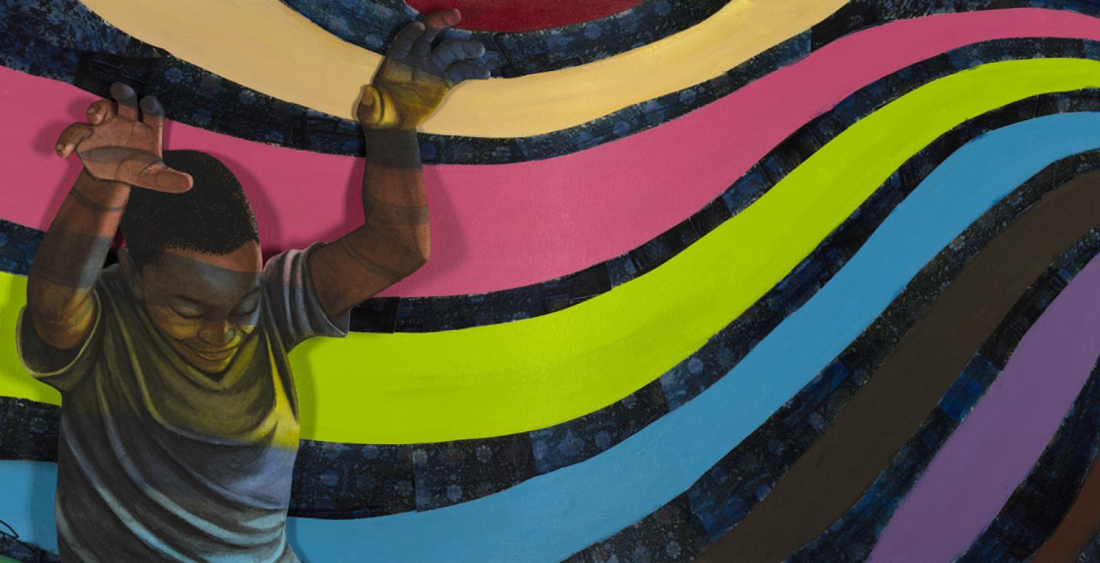 An African American boy stands in front of  rainbow with his hands up. 