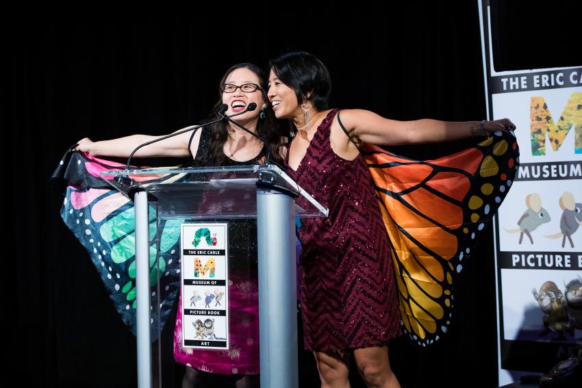 Grace Lin and Alvina Ling stand at a podium wearing butterfly wings as they host The Carle Honors in 2019 at Guastavino's