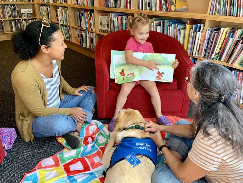 Child sitting in chair showing picture book to reading buddy dog.