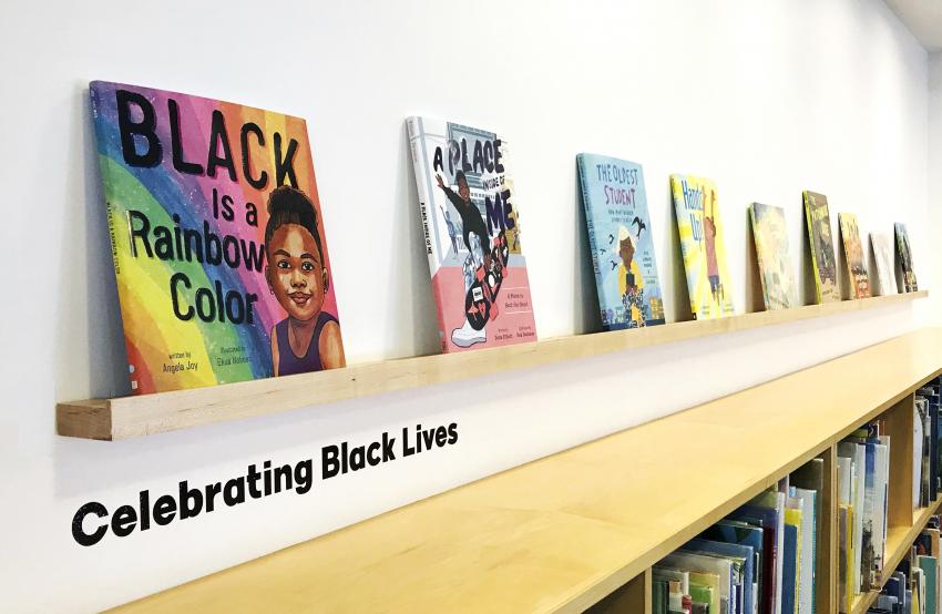Colorful books sit on a wood ledge against a white wall. The black text on the wall reads "Celebrating Black Lives."