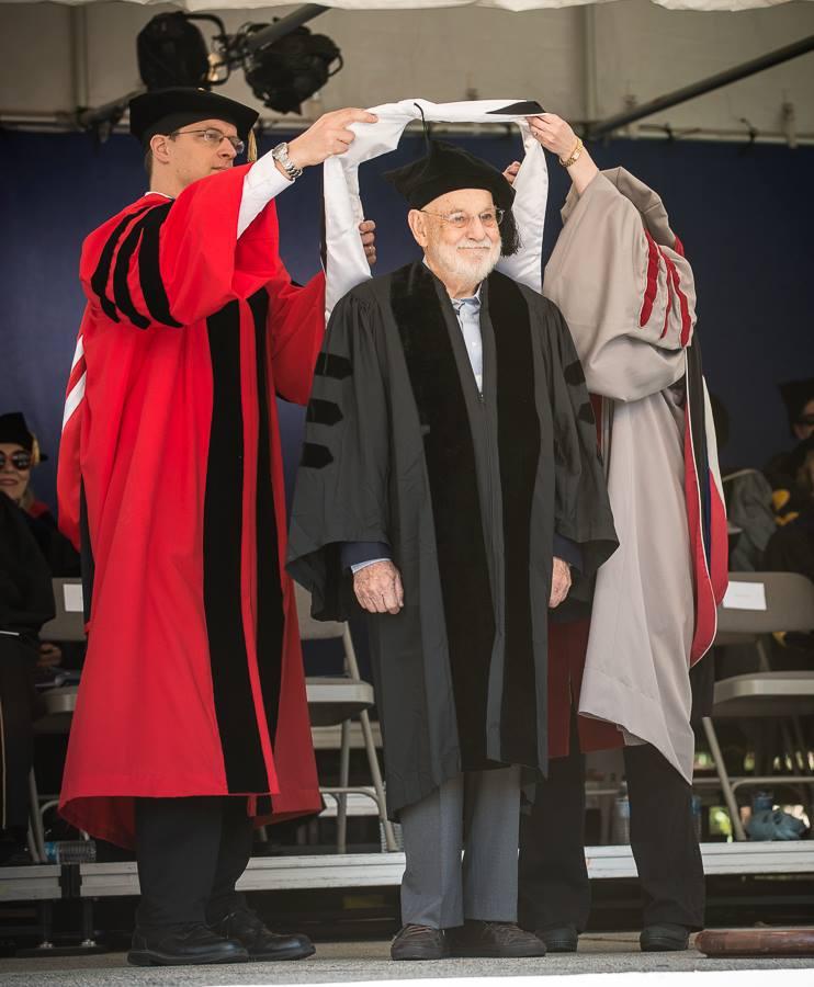 Eric Carle wearing black cap and gown with two people in red and gray gowns putting a white honorary degree hood over his head