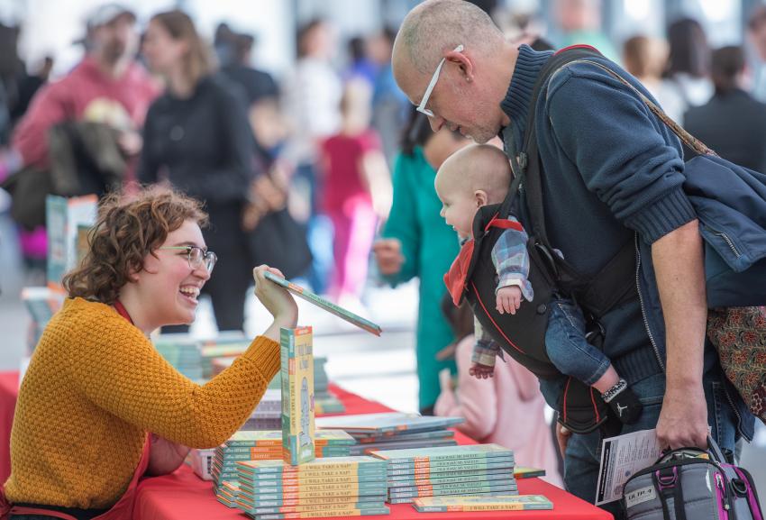 In The Carle's Great Hall, a Museum staff member smiles and shows a book to a baby being carried by a parent