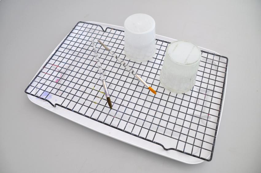 A drying rack made from a lid and a cookie drying sheet, with two brushes and two cups sitting on top drying.