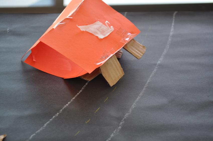An orange paper sculpture on a drawn road with small cardboard wheels.