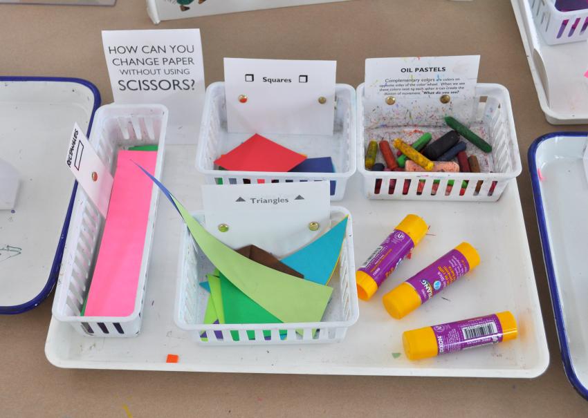 A materials tray including different colors and shapes of paper, oil pastels, and glue sticks.