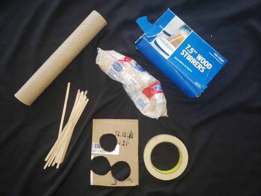 A paper towel roll, wooden stirrers, masking tape, a bag of barley, and a piece of cardboard. 