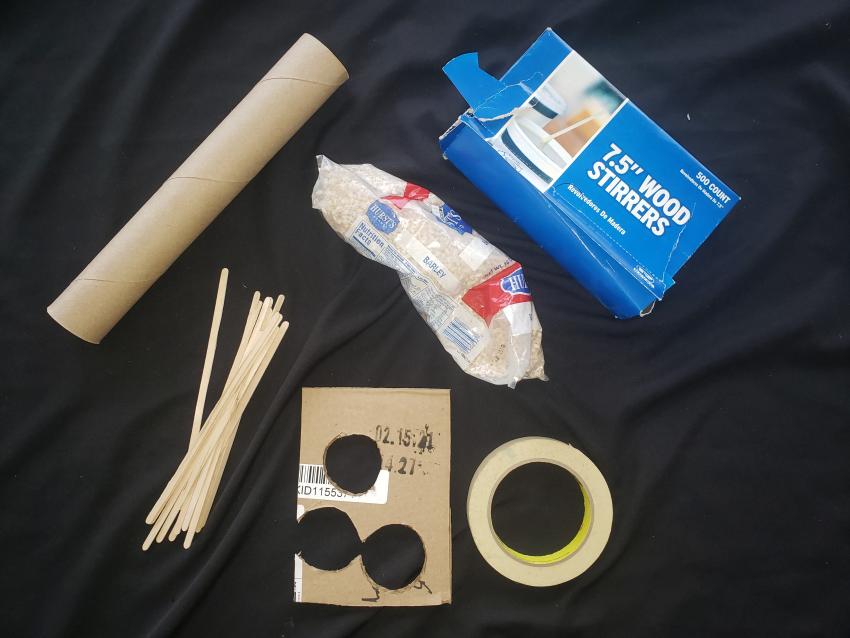 A paper towel roll, wooden stirrers, masking tape, a bag of barley, and a piece of cardboard.