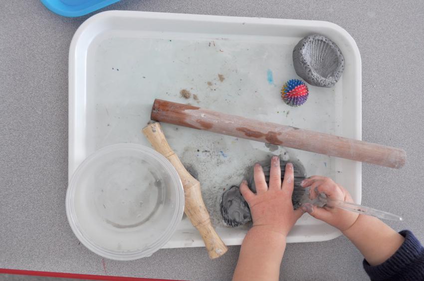 Toddler hands manipulating grey clay on a tray next to a cup of water and several rolling pins.