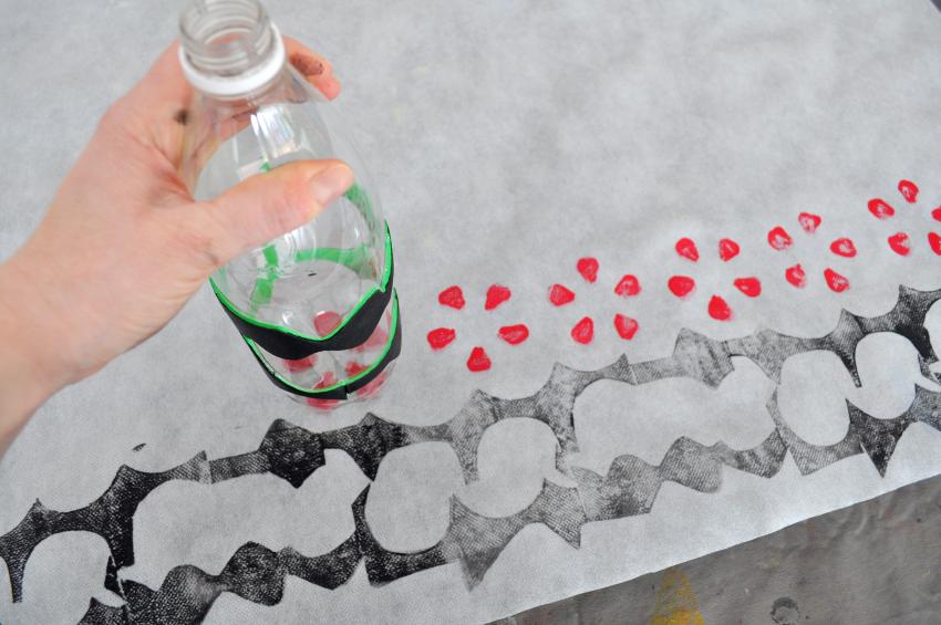 A hand presses a large plastic bottle that was dipped in red paint on white paper, revealing a flower shape stamp.