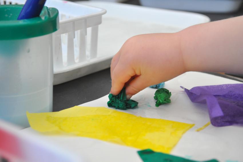 A small hand gently places a tissue paper ball onto a paper with glue on it.