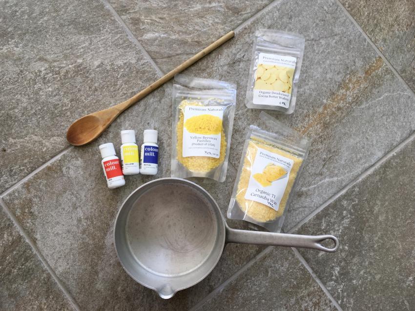 Ingredients to make crayons including a silver pot, wooden spoon, food coloring bottles, and packages of beeswax, carnauba wax, and cocoa butter.